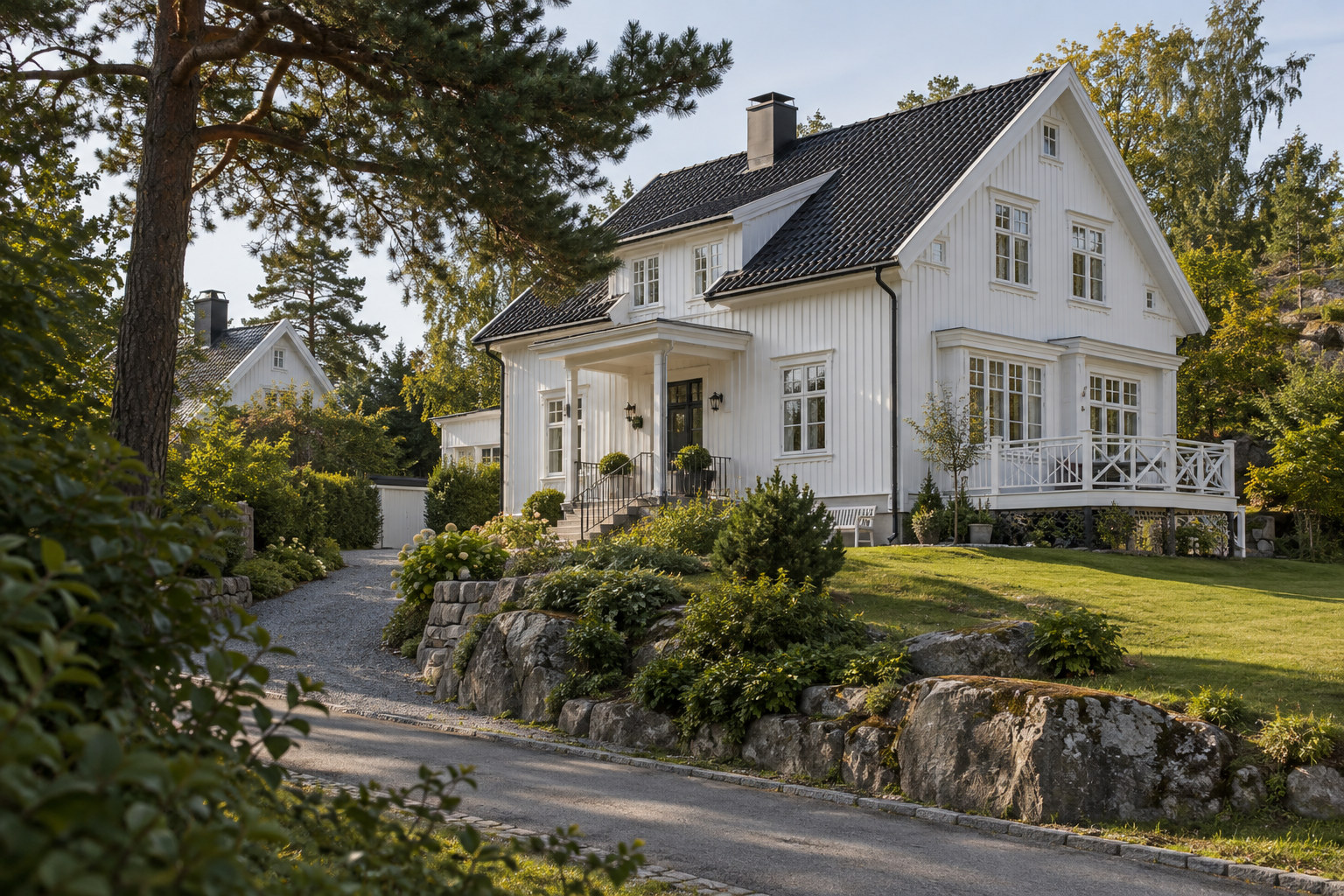 Hvit trevilla med mørkt tak, veranda og hage med furutrær og berg