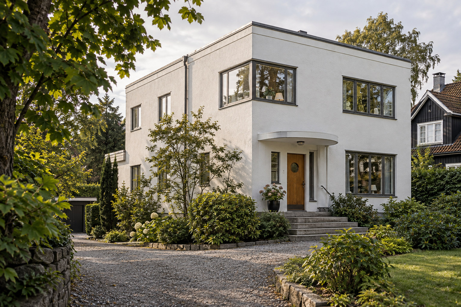 Hvit funkisvilla med mørke vindusrammer, inngangsdør i tre og frodig hage