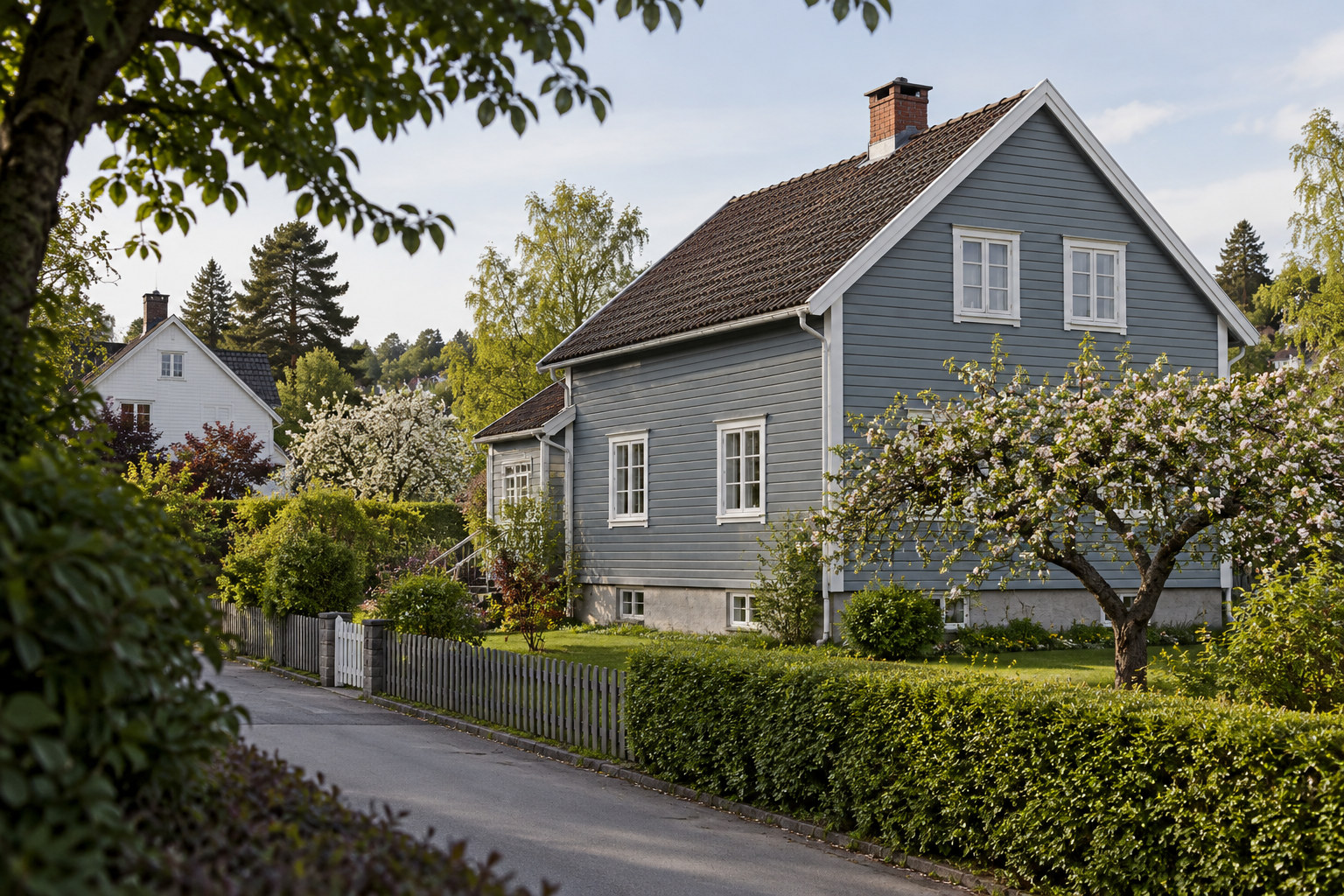 Blågrå trevilla med hvite vindusrammer, skråtak og frodig hage langs en boligvei