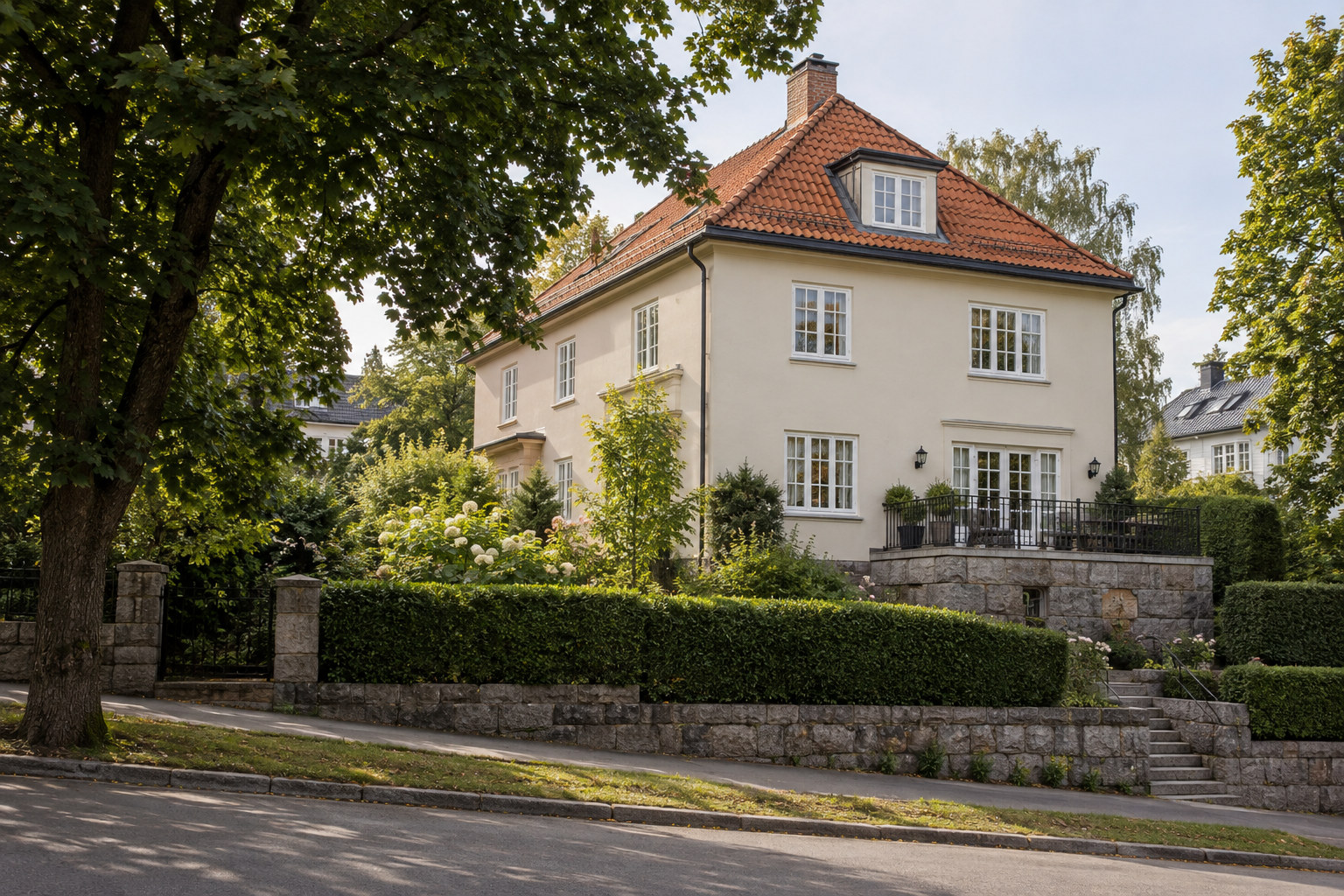 Lys pusset villa med rødt tegltak, sprossevinduer og frodig hage bak en steinmur